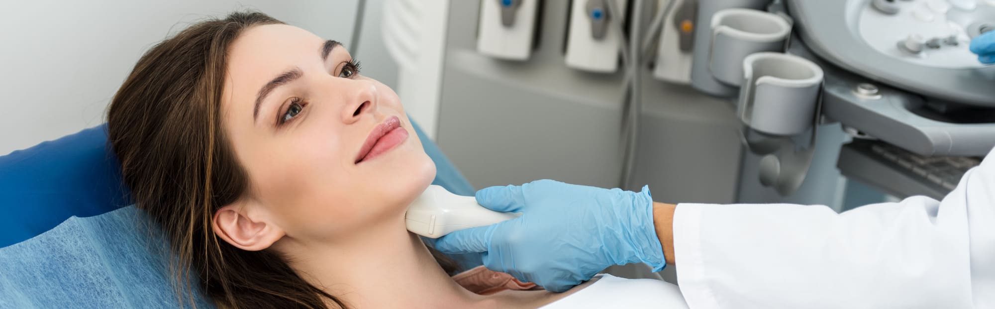 A woman lying on a medical chair receives an ultrasound on her neck. The doctor, wearing blue gloves, uses a handheld device. The room is clinical and calm.